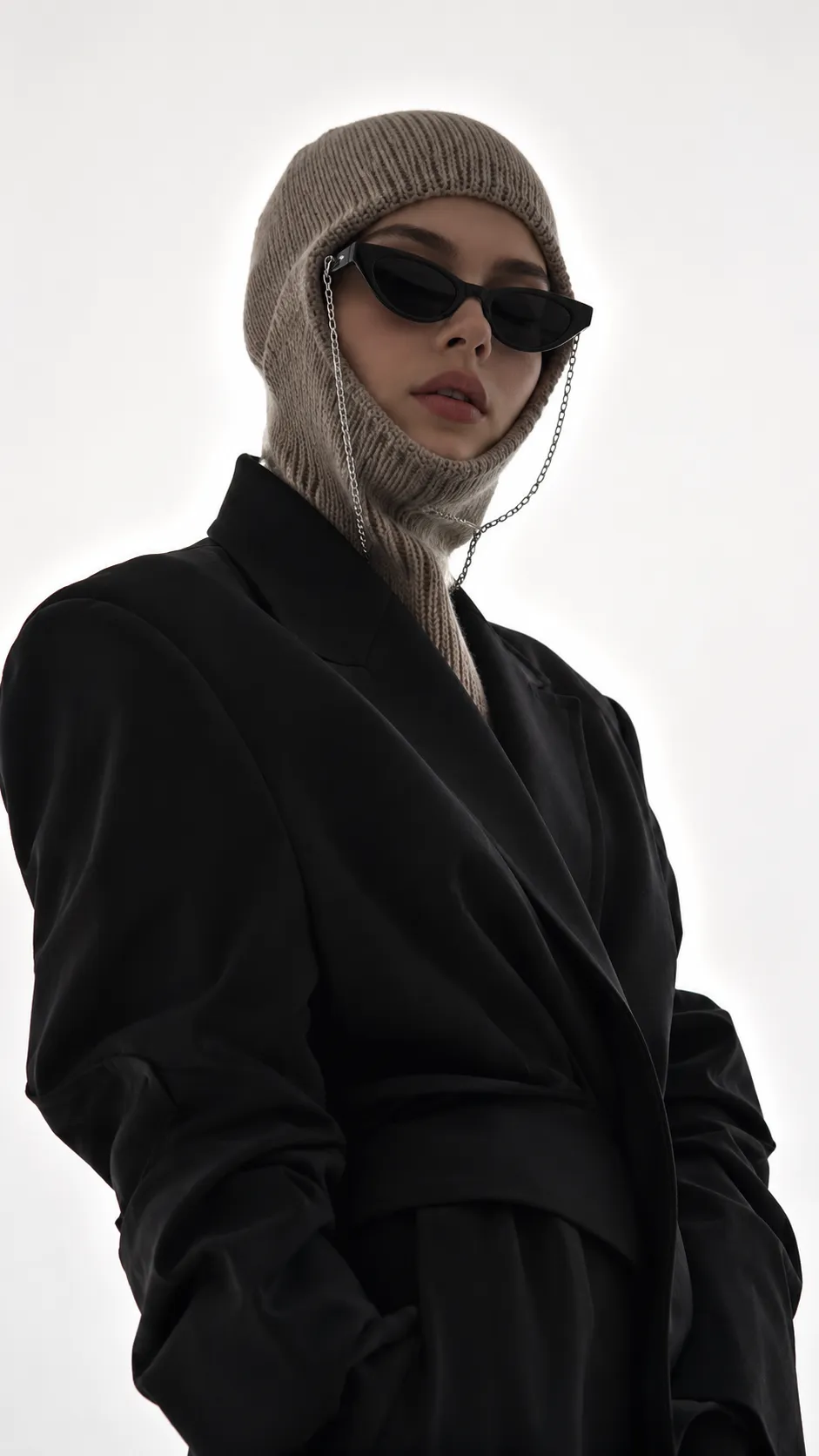 Minimal high-fashion portrait of a stylish young woman wearing a knitted balaclava and black sunglasses against a bright white background.