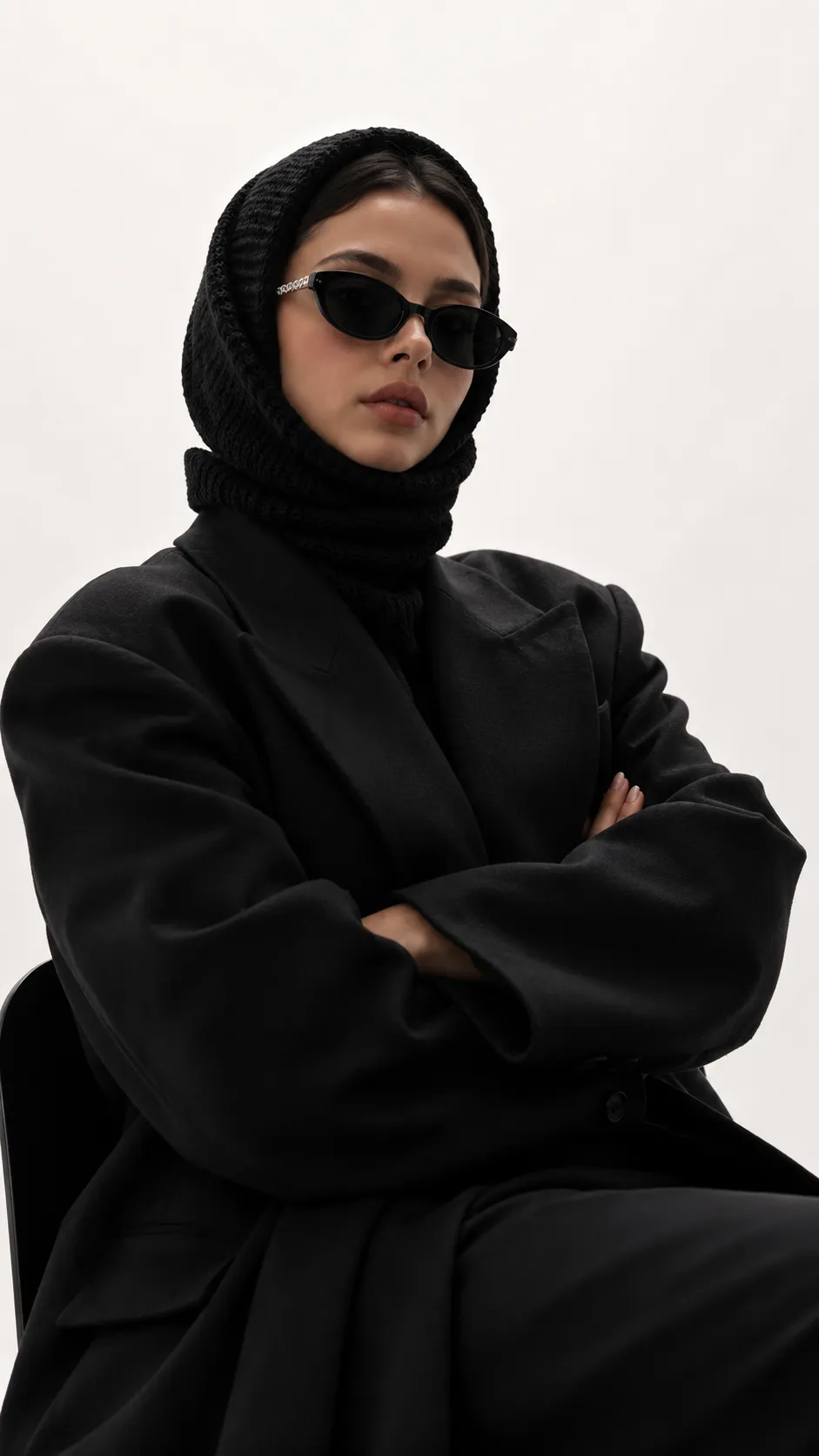 Luxury editorial fashion portrait of a woman wearing a knitted hood and modern sunglasses in a minimalist white studio.